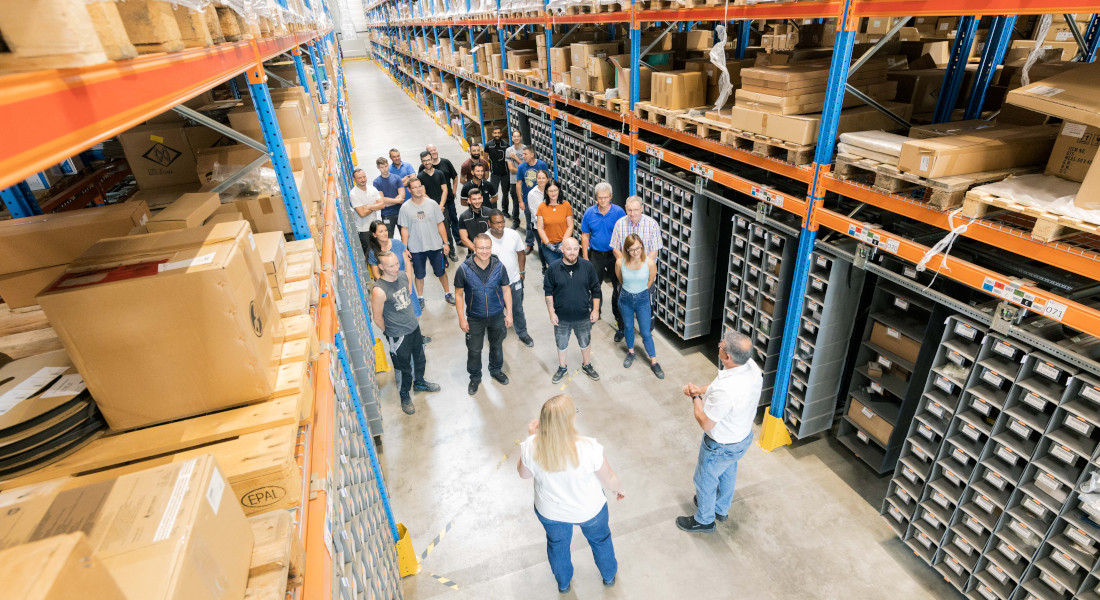 Teammeeting im Lager von EFB-Elektronik mit Mitarbeitenden aus Logistik und Verwaltung zwischen Hochregalen