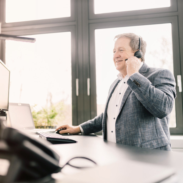 Geschäftsmann im modernen Büro telefoniert mit Headset und arbeitet am Computer vor großen Fenstern. Geschäftsmann im modernen Büro telefoniert mit Headset und arbeitet am Computer vor großen Fenstern.
