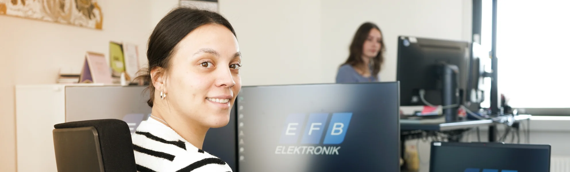 Mitarbeiterin von EFB-Elektronik sitzt lächelnd an einem Büroarbeitsplatz mit mehreren Monitoren