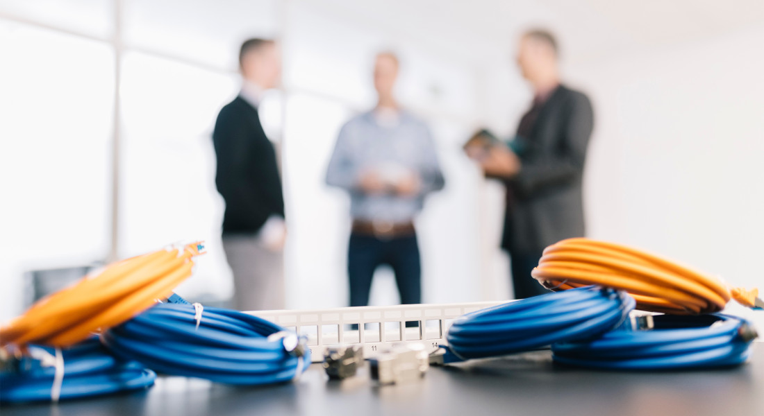 Orange und blaue Netzwerkkabel sowie Patchpanel im Vordergrund, drei Personen im Hintergrund.