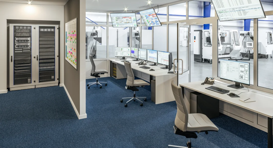 Leitstand einer industriellen Produktion mit Monitoren, Netzwerkschrank und vernetzten Maschinen für die Signal- und Datenübertragung.