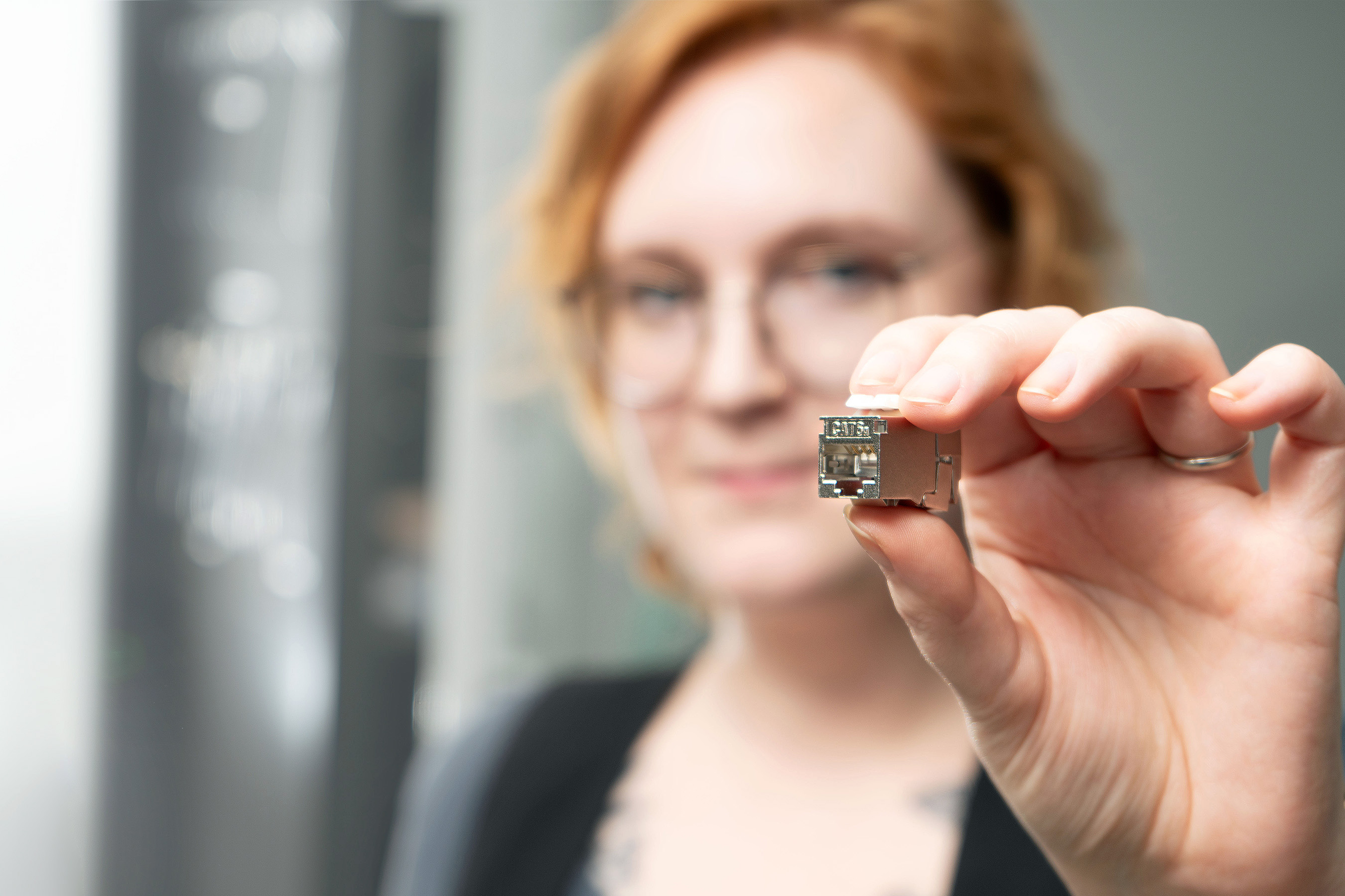 Red-haired woman with round glasses holds RJ45 Keystone Slimline/ Shortbody Cat.6A in her hand