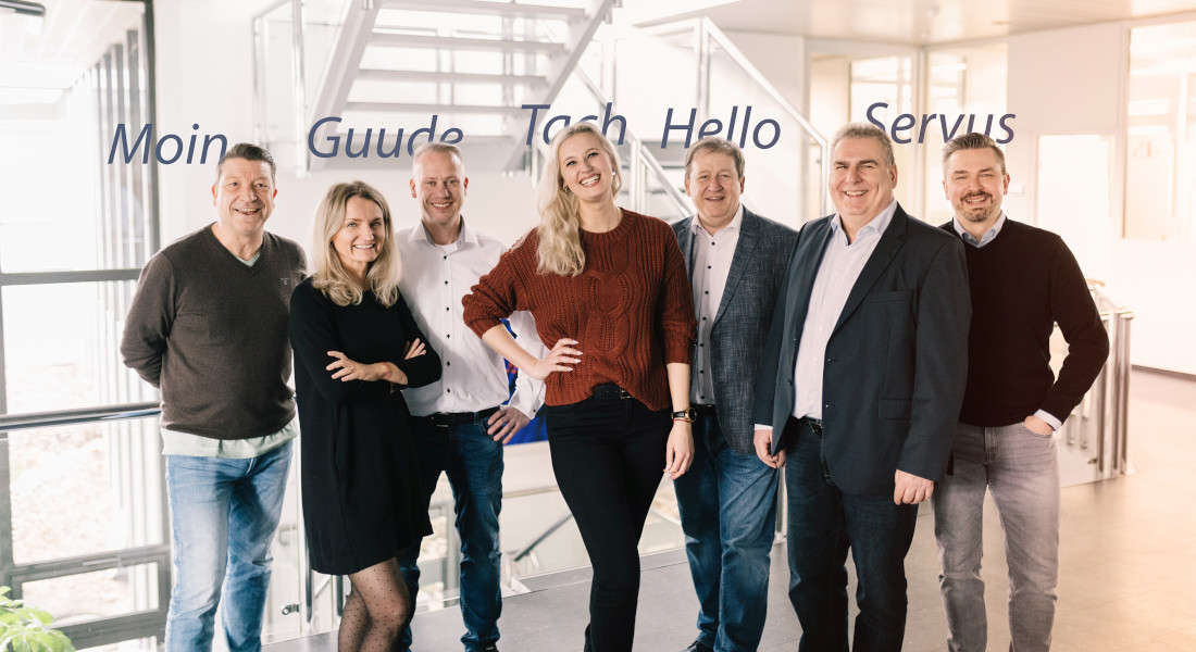 Employees of EFB-Elektronik stand in the entrance area. Above them, the regional greetings "Moin", "Servus", "Guude", "Tach", "Hello" are inserted as text.
