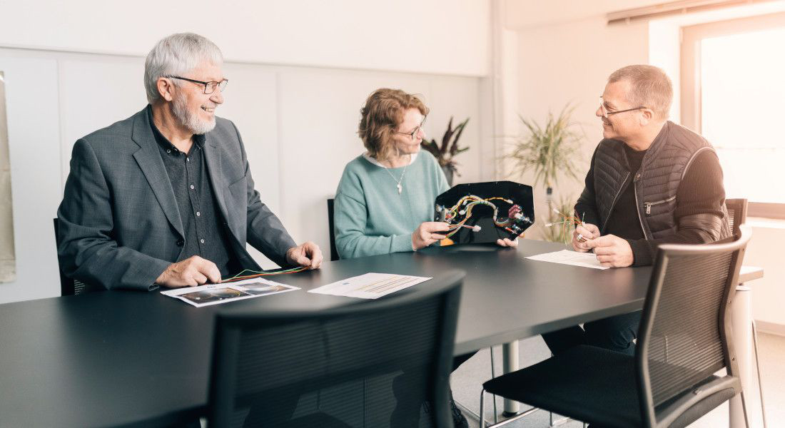 Drei Personen bei einem Gespräch am Tisch, eine Frau hält ein technisches Bauteil.