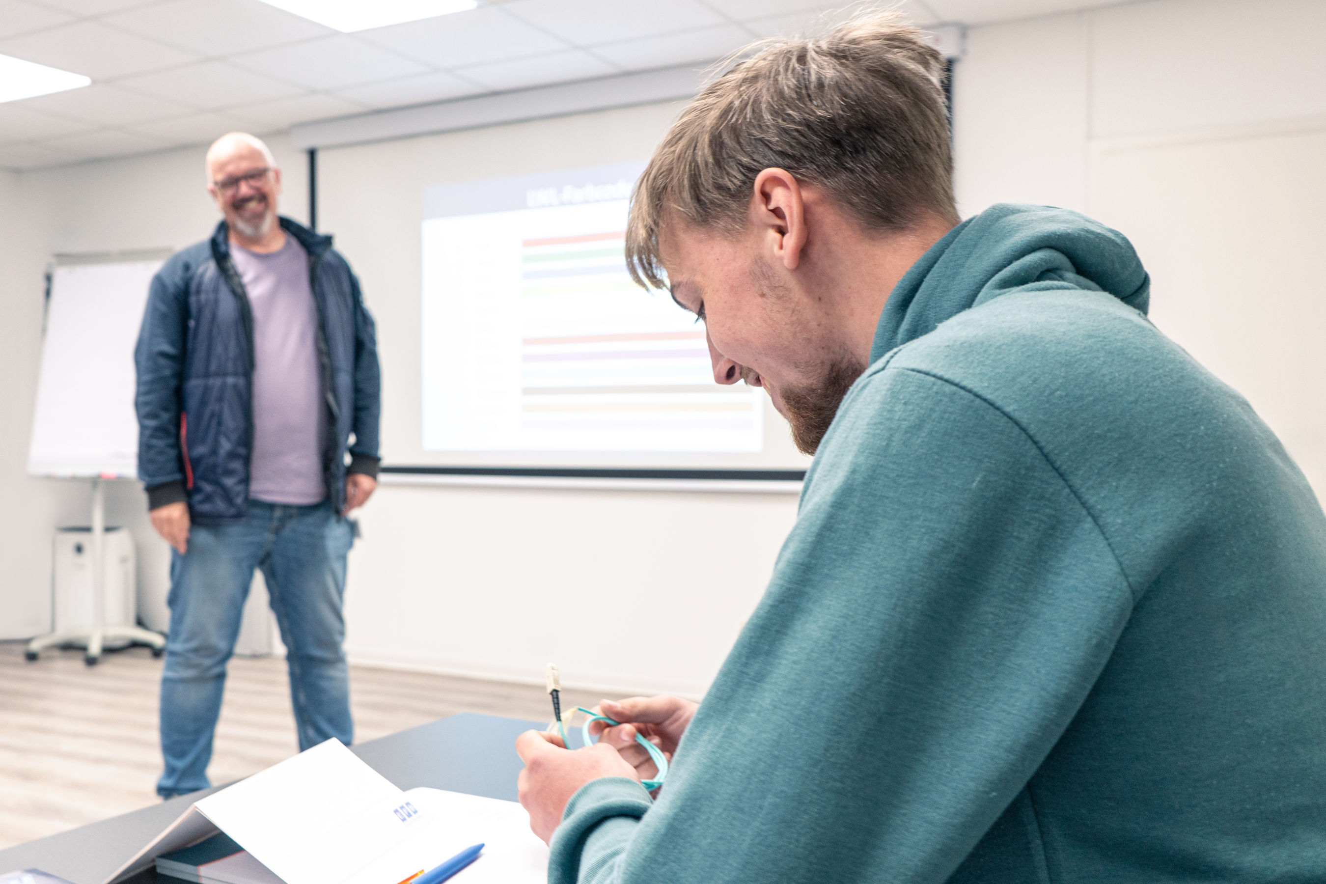 Dozent steht vorne in einem Schulungsraum und hält eine Präsentation, während ein Teilnehmer Notizen macht