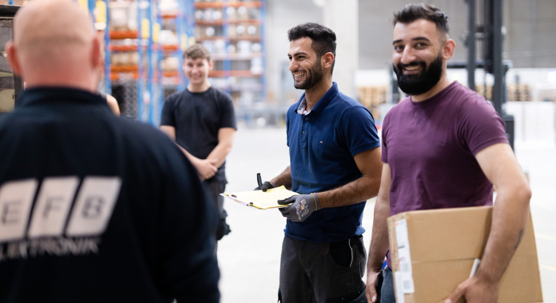 Teammeeting im Lager von EFB-Elektronik mit Mitarbeitenden im Austausch und Paketübergabe in freundlicher Atmosphäre
