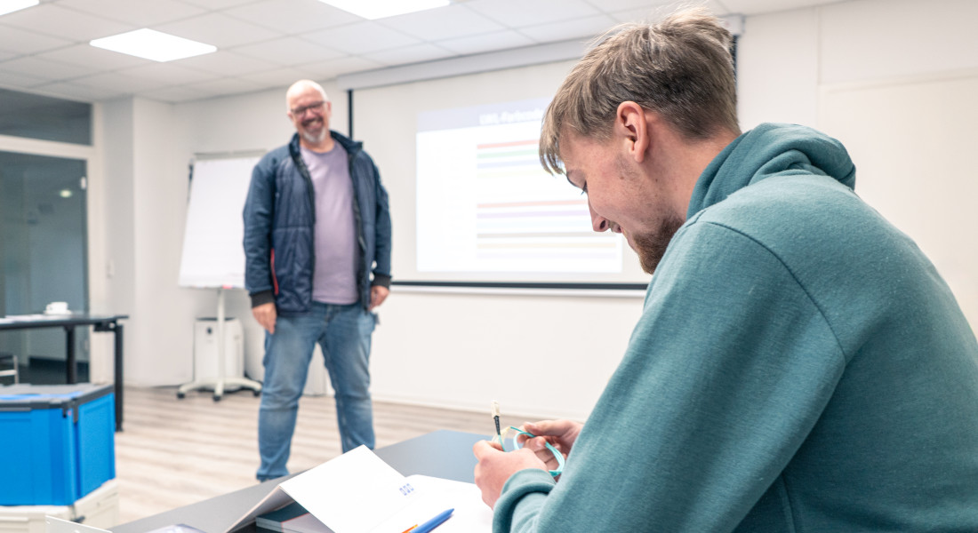 Dozent steht vorne in einem Schulungsraum und hält eine Präsentation, während ein Teilnehmer Notizen macht