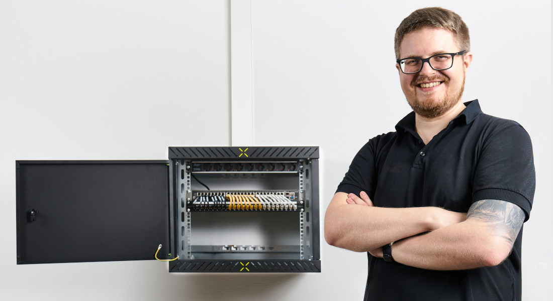Installateur vor montiertem X-Rack 19"-Wandgehäuse an der Wand – Tür geöffnet, Komponenten übersichtlich installiert, ideal für strukturierte Netzwerkverkabelung und schnellen Servicezugang.